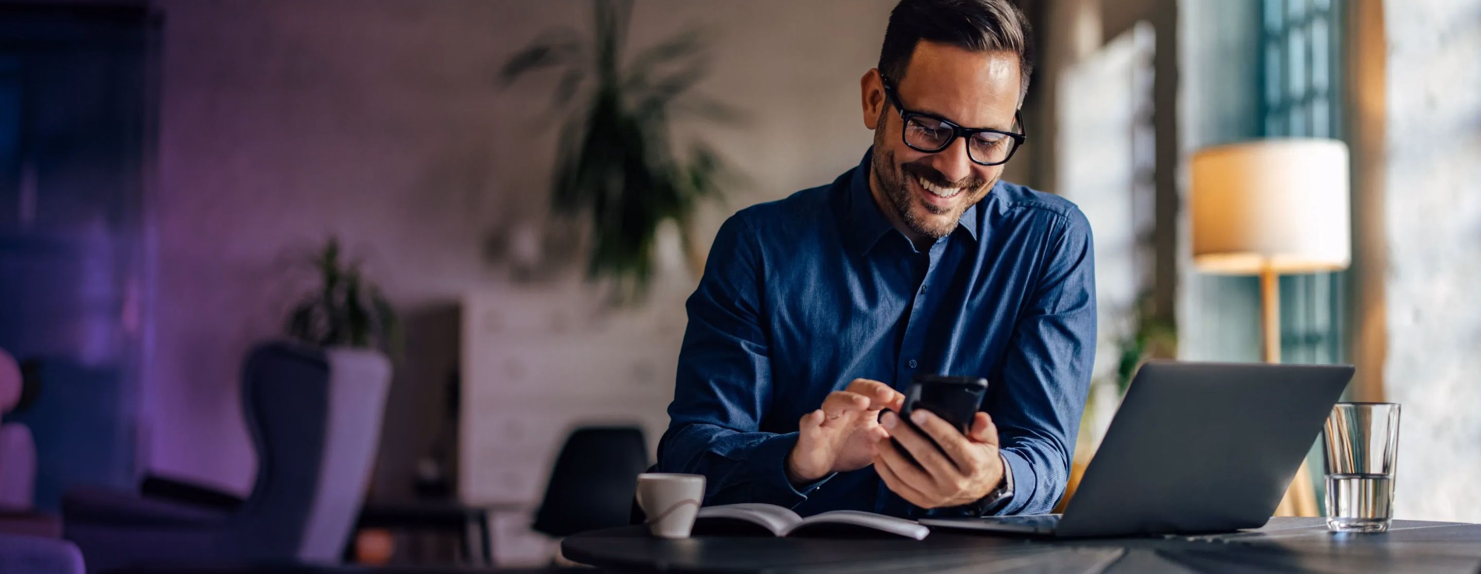 Man smiling at his phone while working at a desk with a laptop
